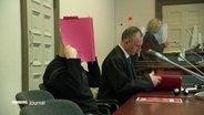 Ein Angeklagter hält sich im Gerichtssaal eine rote Mappe vor das Gesicht, neben ihm sitzt sein Verteidiger. © Screenshot 