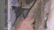 Eine Hand legt eine unter Weißer Farbe verstecktes Wandgemälde mit einem Kratzer frei. © Screenshot 