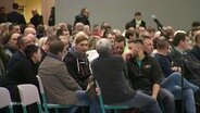 Viele Menschen sitzen bei einer Bürgerversammlung in Stuhlreihen in einer Turnhalle. © Screenshot 