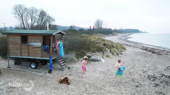 Ein kleines Häuschen auf Rädern steht an einem Strandabschnitt. Drei Menschen in Badekleidung gehen Richtung Wasser. © Screenshot 