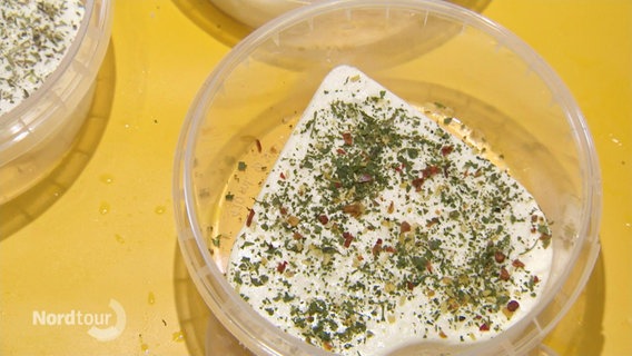 Ein herzförmiger und mit Kräutern bestreuter Käse liegt in einer Plastikbox. © Screenshot 