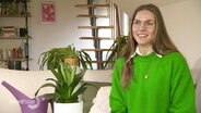 Eine junge Frau sitzt auf einem Sofa neben Grünpflanzen und einer Gießkanne. © Screenshot 