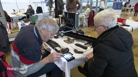 Zwei Menschen sitzen an einem Tisch und untersuchen die darauf liegenden Elektrogeräte. © Screenshot 