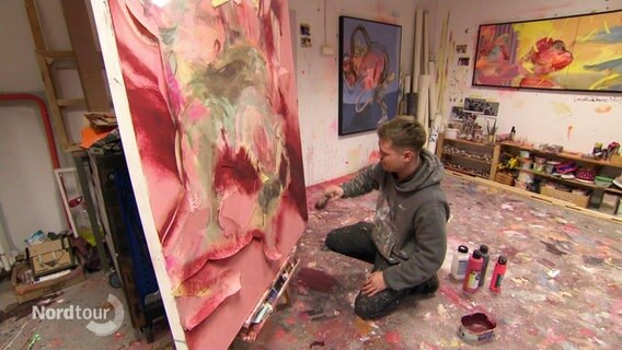 Ein junger Mann arbeitet in einem Artelier an einem großen Gemälde. © Screenshot 