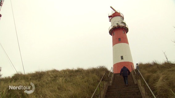 Ein Mann geht eine Treppe hinauf zu einem rot-weiß gestreiften Leuchtturm. © Screenshot 