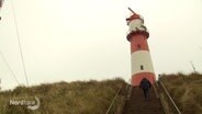 Ein Mann geht eine Treppe hinauf zu einem rot-weiß gestreiften Leuchtturm. © Screenshot 