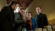 Vier Menschen stehen hinter einem Stapel Bücher in einer Kirche und betrachten erschüttert ein Bild auf einen Fotoapparat. © Screenshot 