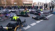 Viele Menschen liegen bei einer Mahnwache auf dem Rücken auf einer Straßenkreuzung. Neben ihnen liegen Fahrräder. Im Hintergrund stehen weitere Personen. © Screenshot 