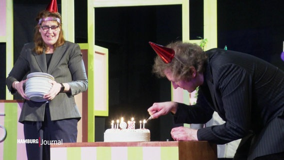 Zwei Schauspielende auf einer Bühne. Der eine zündet Kerzen auf einer Torte an, die andere hält einen Stapel Teller. © Screenshot 