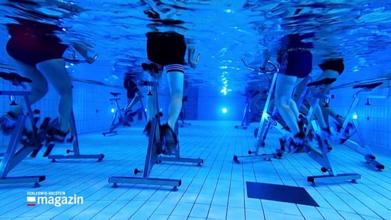 Menschen sitzen auf Trimm-Räder, die unter Wasser in einem Schwimmbecken stehen. © Screenshot 