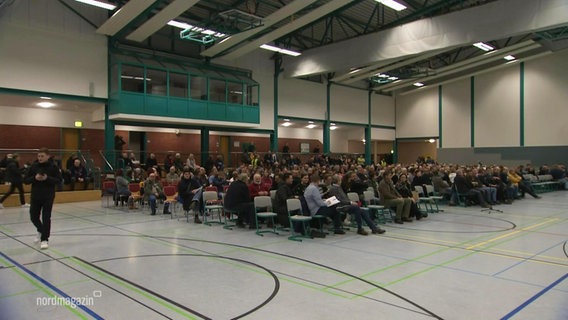 Uphaler sitzen zusammen in einer Turnhalle und nehmen an einer Veranstaltung zum Bau eines Containerdorfes für Geflüchtete teil. © Screenshot 