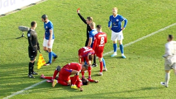 Ein Schiedsrichter zwischen Fußballspielenden hält eine rote Karte hoch. © Screenshot 