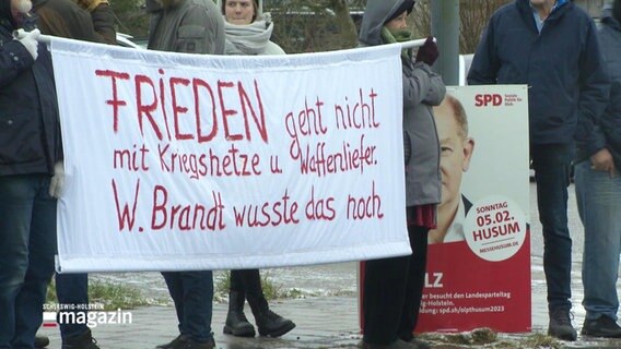 demonstrierende Menschen halten ein Banner mit der Aufschrift: "Frieden geht nicht mit Kriegshetze und Waffenlieferung, W. Brand wusste das noch" © Screenshot 