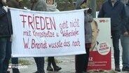 demonstrierende Menschen halten ein Banner mit der Aufschrift: "Frieden geht nicht mit Kriegshetze und Waffenlieferung, W. Brand wusste das noch" © Screenshot 