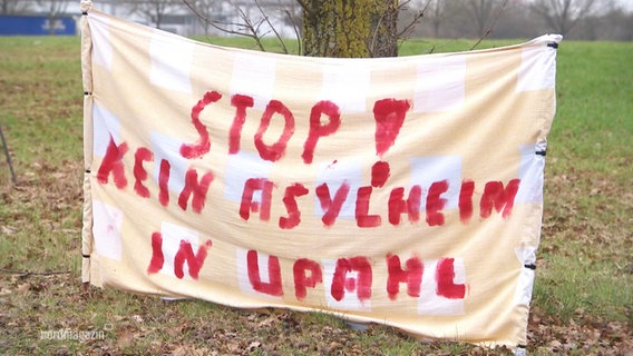 An einem Baum vor einem Feld ist ein Bettlaken aufgehängt auf der in großer Schrift mit roter Farbe geschrieben steht: "Stop! Kein Asylheim in Upahl". © Screenshot 