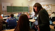 Eine Lehrerin steht neben einer ihrer Schülerinnen im Klassenzimmer. © Screenshot 