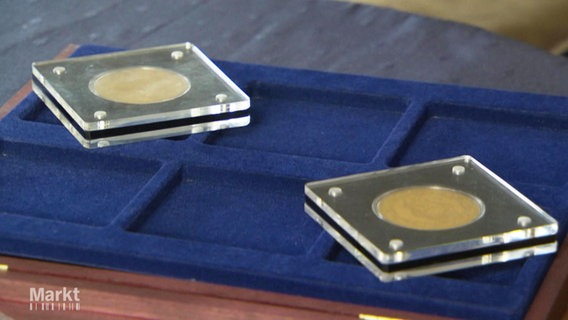 Auf einem blau-samtenen Behältnis liegen zwei in Plastikscheiben geschütze Goldmünzen. © Screenshot 