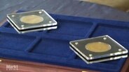 Auf einem blau-samtenen Behältnis liegen zwei in Plastikscheiben geschütze Goldmünzen. © Screenshot 