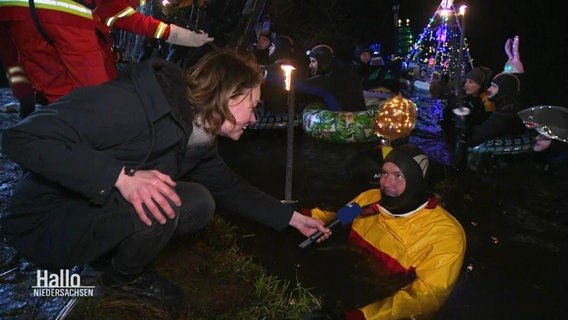 Eine Reporterin interviewt einen Rettungsschwimmer vom Ufer aus. Der Schwimmer ist im Wasser, hat einen Anzug an und eine Fackel in der Hand. © Screenshot 