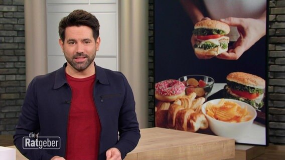 Der Mann steht im Studio, hinter ihm ist auf einem Bild ein Burger abgebildet. © Screenshot 