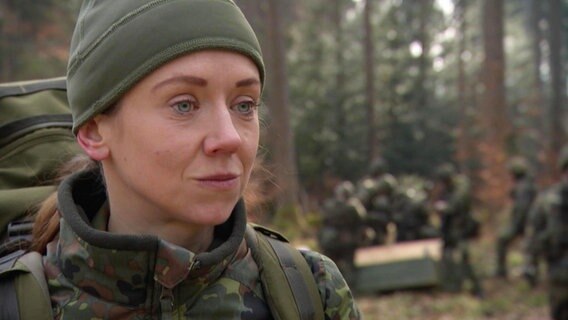 Tina Behnke steht in Tarnkleidung mit einem Rucksack in einem Wald. © Screenshot 