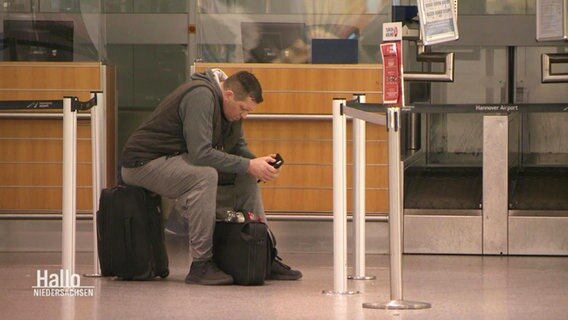 Ein Mann sitzt vor einem Flughafenschalter auf seinem Koffer und schaut auf sein Handy. © Screenshot 