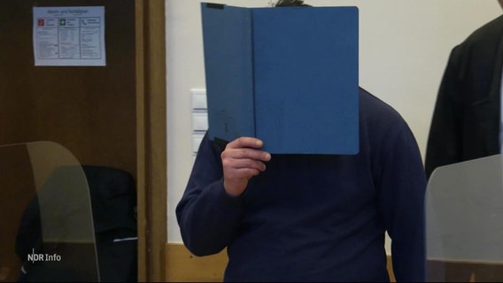 Ein Angeklagter hält sich eine Aktenordner vor das Gesicht im Gerichtssaal. © Screenshot 