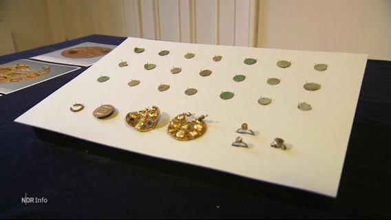 Auf einer weißen Fläche sind mehrere Goldmünzen und Broschen drapiert. © Screenshot 