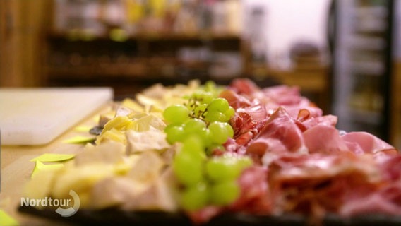 Auf einem längerem Brett sind mehrere Lagen Käse, Schinken und Trauben in Reihen aufgetan. © Screenshot 