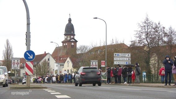 An einer Hauptstraße stehen viele Menschen in einer Menschenkette am Straßenrand und halten sich an den Händen. © Screenshot 