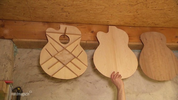 An einem Deckenbalken hängen mehrere Holzsvorschnitte eines Gitarrenklangkörpers. Eine Hand greift von nach einem von ihnen. © Screenshot 