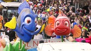 Zwei größere Fischfiguren im Zeichentrickstil sind auf einem Wagen bei einem Karnevalsumzug montiert. Im Hintergrund: zahlreiche Menschen stehen am Straßenrand. © Screenshot 