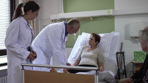 Ein Arzt und eine Ärztin stehen am Bett einer Frau und reden mit ihr. © Screenshot 