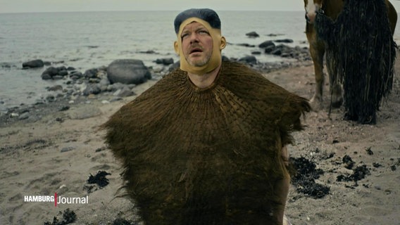 Porky von Deichkind kniet im neuen Musikvideo zum neuen Album mit einem Bastrock um den Hals an einem Strand und schaut gen Himmel an einem trüben Tag. © Screenshot 