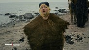 Porky von Deichkind kniet im neuen Musikvideo zum neuen Album mit einem Bastrock um den Hals an einem Strand und schaut gen Himmel an einem trüben Tag. © Screenshot 