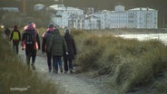 Mehrere Menschen laufen einen kleinen Wanderweg in einer Stranddüne entlang. © Screenshot 