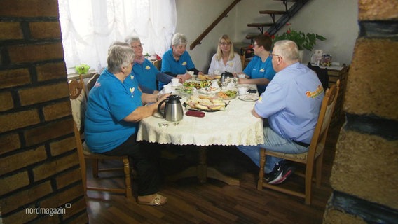 Mehrere ältere Damen in blauen T-Shirts sitzen mit zwei älteren Personen in normaler Kleidung an einer größeren Kaffeetafel. © Screenshot 