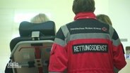Von hinten sind ein Mann in der Berufskleidung des Deutschen Roten Kreuzes zu sehen; auf der Jacke steht in Großbuchstaben "Rettungsdienst" auf dem Rücken. Er schiebt eine Liege mit einem Patienten. © Screenshot 