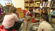 Vier Kinder und zwei erwachsene Frauen sitzen auf dem Fußboden und spielen, hinter ihnen steht ein Regal mit vielen Brettspielen und anderen Spielsachen. © Screenshot 