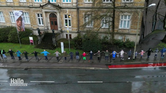 Blick aus der Vogelperspektive: Mehrere Menschen stehen Hand in Hand in einer langen Menschenkette auf dem Bürgersteig einer größeren Hauptstraße. © Screenshot 