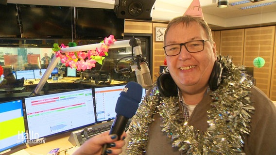 Moderator Michael Thürnau steht im Radiostudio, trägt ein Partyhütchen und grinst in die Kamera. © Screenshot 