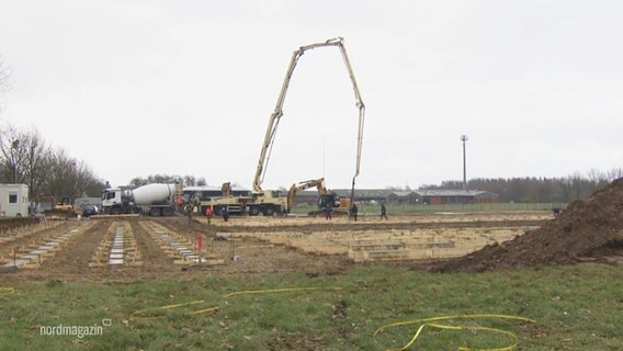 Auf einem größeren Feld wird mit einem großen Kranarm aus einem Betonmischer das Fundament für eine zukünftige Unterkunft für Geflüchtete gelegt. © Screenshot 