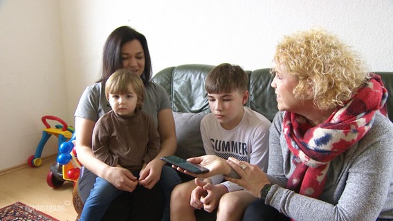 Eine Mutter mit ihren zwei jüngeren Söhnen sitzt mit einer Reporterin auf einem Sofa in einer Wohnung. © Screenshot 