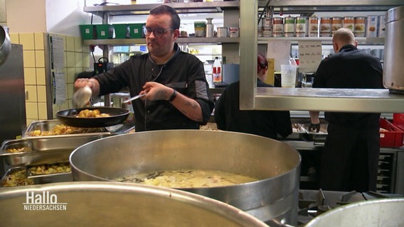 Ein Mann füllt in einer Großküche Essen aus einem großen Topf in eine Pfanne. © Screenshot 