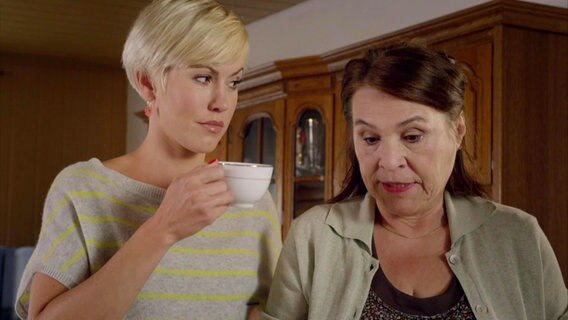 Die Protagonistin Klara und eine ältere Dame nebeneinander in einem Wohnzimmer; Klara hält eine Teetasse in der Hand und schaut die Dame an. © Screenshot 