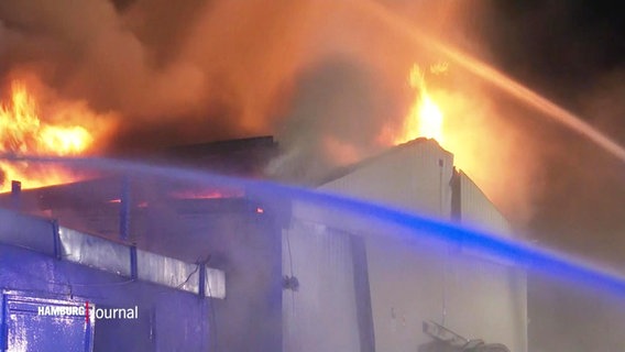 Das Dach einer Lagerhalle steht in Flammen, man sieht zwei Löschwasserstrahle der Feuerwehr. © Screenshot 