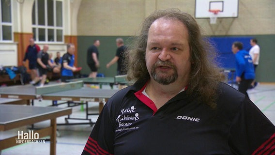 Ein Mann im Interview in einer Sporthalle, im Hintergrund spielen andere Tischtennis. © Screenshot 