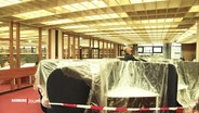 Mit Plane abgedecktes Mobiliar in der Staatsbibliothek Hamburg, da es durch das Dach regnet. Ein Mann schaut zur Decke. © Screenshot 