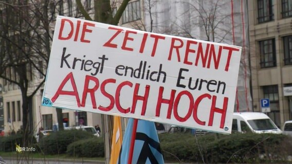 Ein Schild auf einer Demonstration mit der Aufschrift: "DIE ZEIT RENNT KRIEGT ENDLICH EUREN ARSCH HOCH". © Screenshot 
