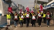Mitarbeitende der HADAG beim Warnstreik an den Landungsbrücken. Sie tragen Schilder mit den Buchstaben H,A,D,A,G und ein Ausrufezeichen. © Screenshot 